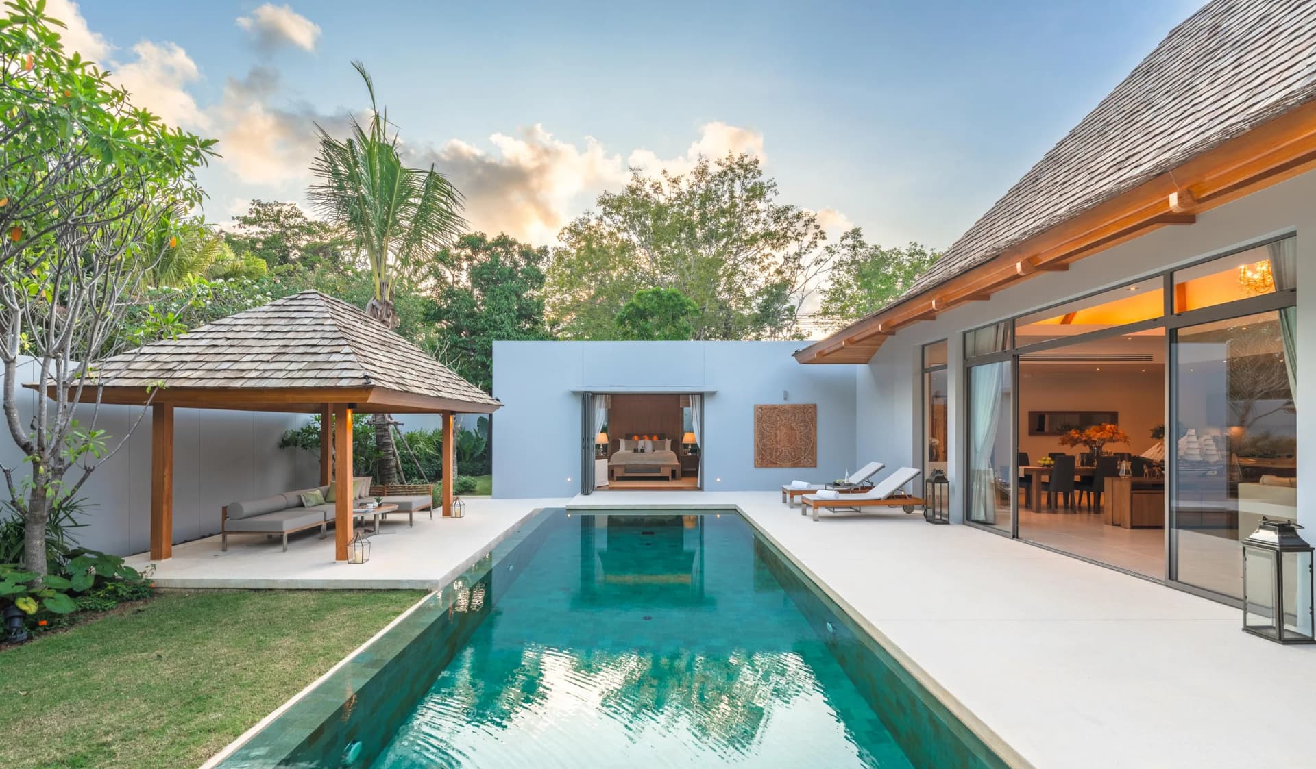 Am Pool einer gebauten Anchan-Villa: eine Holz-Sala mit Daybed, Sonnenliegen und das zur Terrasse offene Wohnzimmer