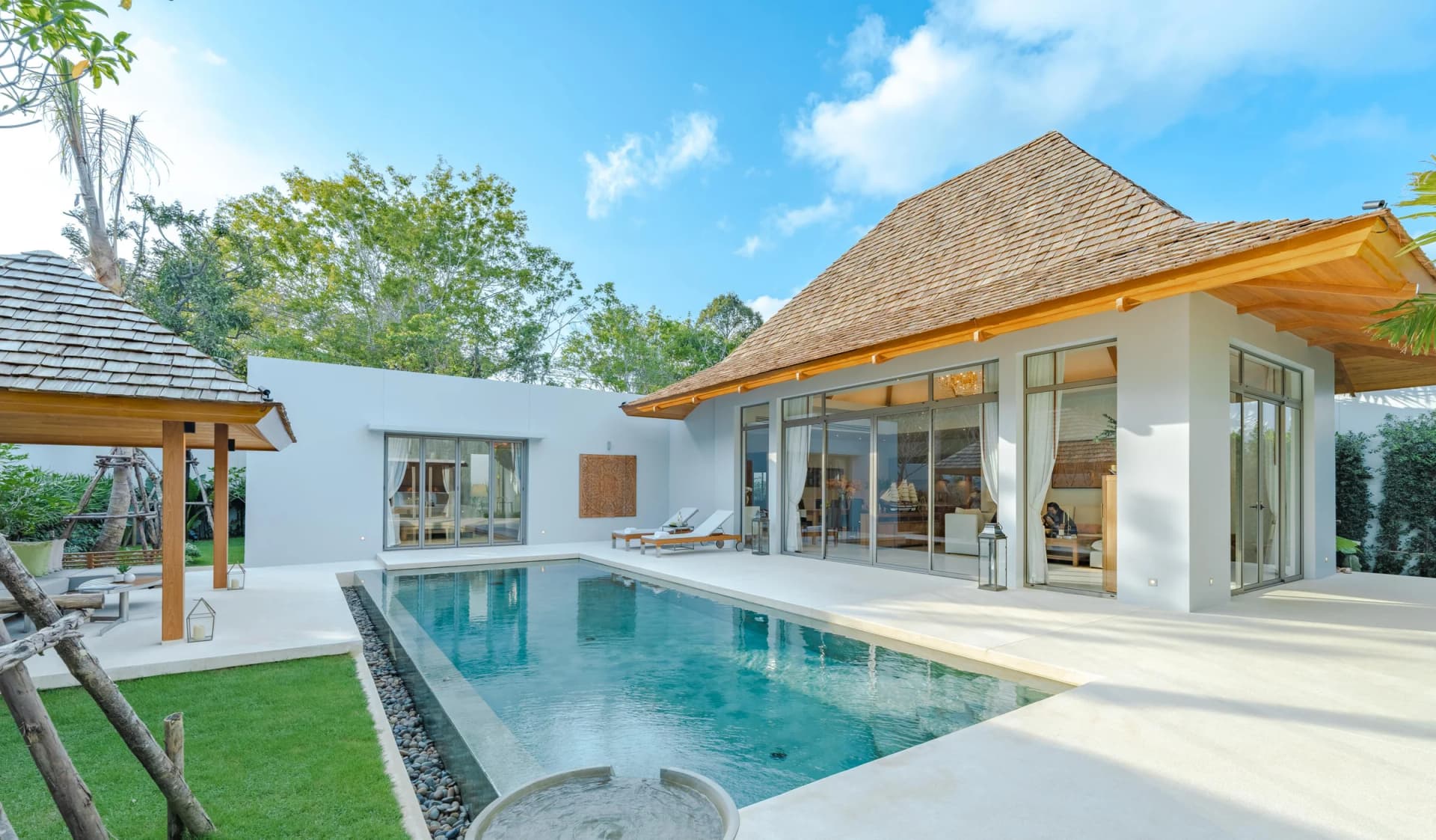 Eine gebaute Anchan-Villa in voller Sonne: weiße Wände, hohe Glastüren unter einem Holzschindeldach, der Pool mit Kieselrand und eine Holz-Sala